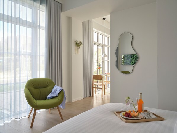 Habitación de hotel con cama, una silla verde, bandeja de desayuno con fruta y jugo, y vista al exterior a través de amplias ventanas.
