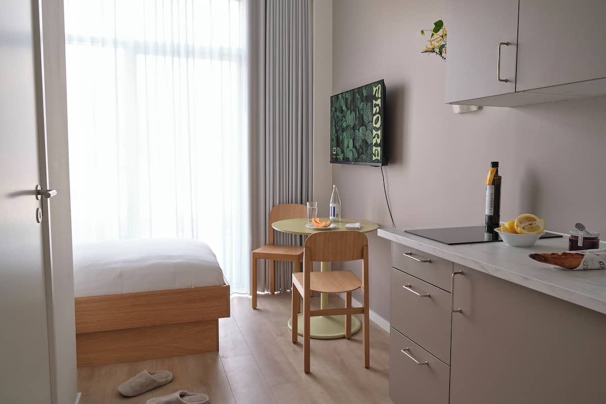 Quarto de hotel com cama, escrivaninha, cadeira, mesinha, televisão e janela com cortinas.