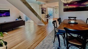 Ein moderner Essbereich mit einem Holztisch, schwarzen Stühlen und einer Chandelüster. Eine Treppe führt in einen weiteren Raum.