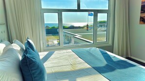 Ein Schlafzimmer mit großem Fenster, einem Bett mit weißer und blauer Bettwäsche sowie Blick auf einen Balkon und einen Außenbereich.