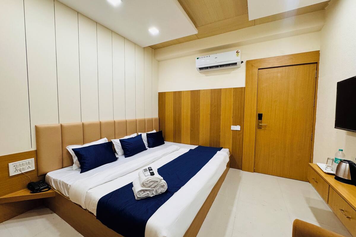 A hotel room with a bed, wooden headboard, a flat-screen TV, a wooden wardrobe, and an air conditioning unit.