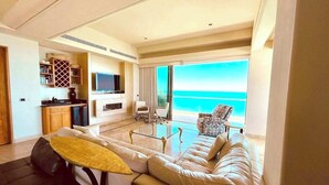 A modern living room with a sofa, a small table, and a TV. There is a kitchen area with cabinets and a stove. The room has large windows with a view of the ocean. - Ocean Wrap Around Oceanfront Balcony at Tessoro (Las Conchas)