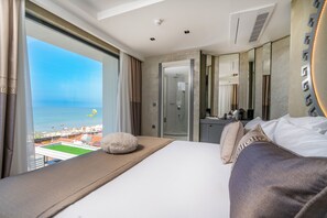 A hotel room with a large window overlooking a beach, a bed with white and brown bedding, a fluffy white pillow, and a mirror on the wall. - Simus Beach Hotel (Manavgat)