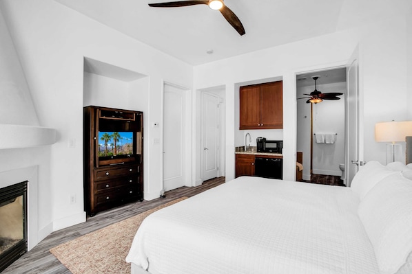 A hotel room with a large bed, a television, a kitchen area, and a ceiling fan.