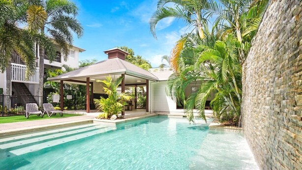 A swimming pool with clear water, surrounded by palm trees and a covered patio area.