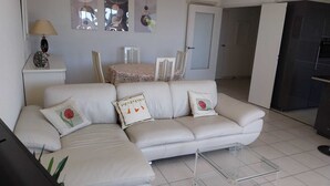 A living room with a white sectional sofa, a small side table with a lamp, and a glass coffee table. There are three wall-mounted mirrors and a door leading to another room. - Location vue mer Saint Raphaël (Saint-Raphaël)