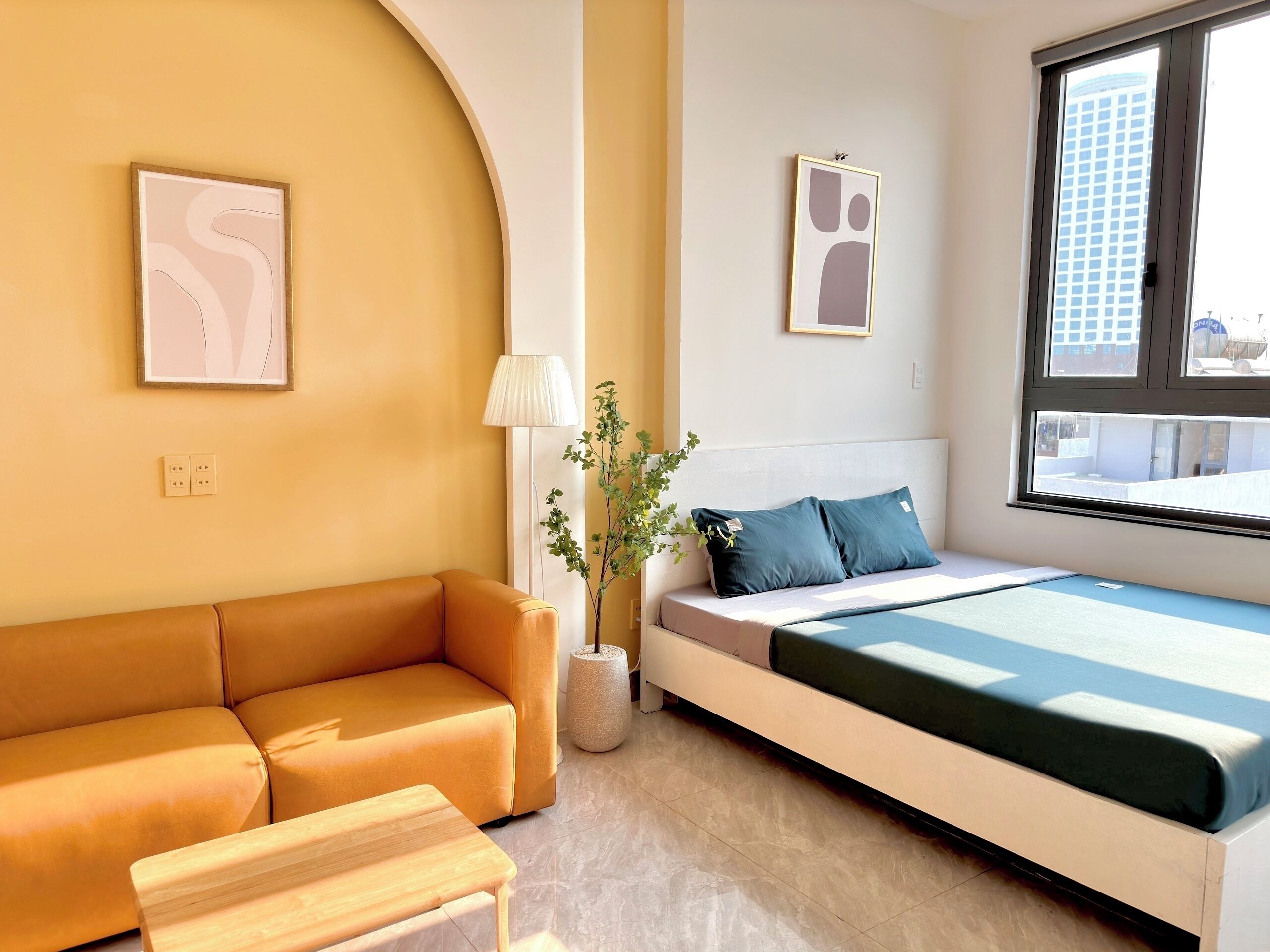 A modern hotel room with a sofa, a bed with a blue bedspread, a potted plant, and two framed artworks on the walls.