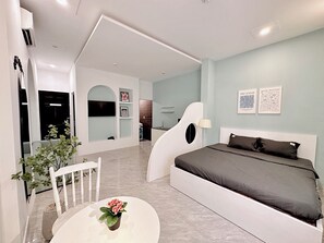 A modern bedroom with a large bed, a small table with a potted plant, and a white chair. There is a bathroom with a shower and a sink visible in the background. - Corgi House Danang (Da Nang)