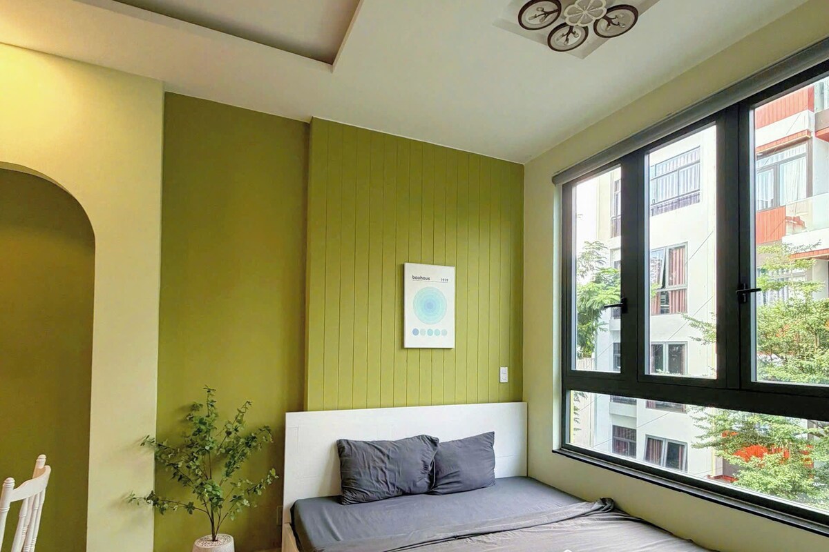 A bedroom with a bed, a potted plant, a white chair, and a window with a view of buildings.