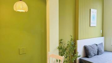 A room with a bed, a table with a chair, a hanging light fixture, a wall-mounted picture, and a potted plant.