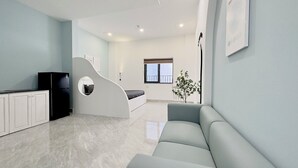 A modern living room with a sofa, a unique white curved structure, a black refrigerator, and a window with blinds. - Corgi House Danang (Da Nang)