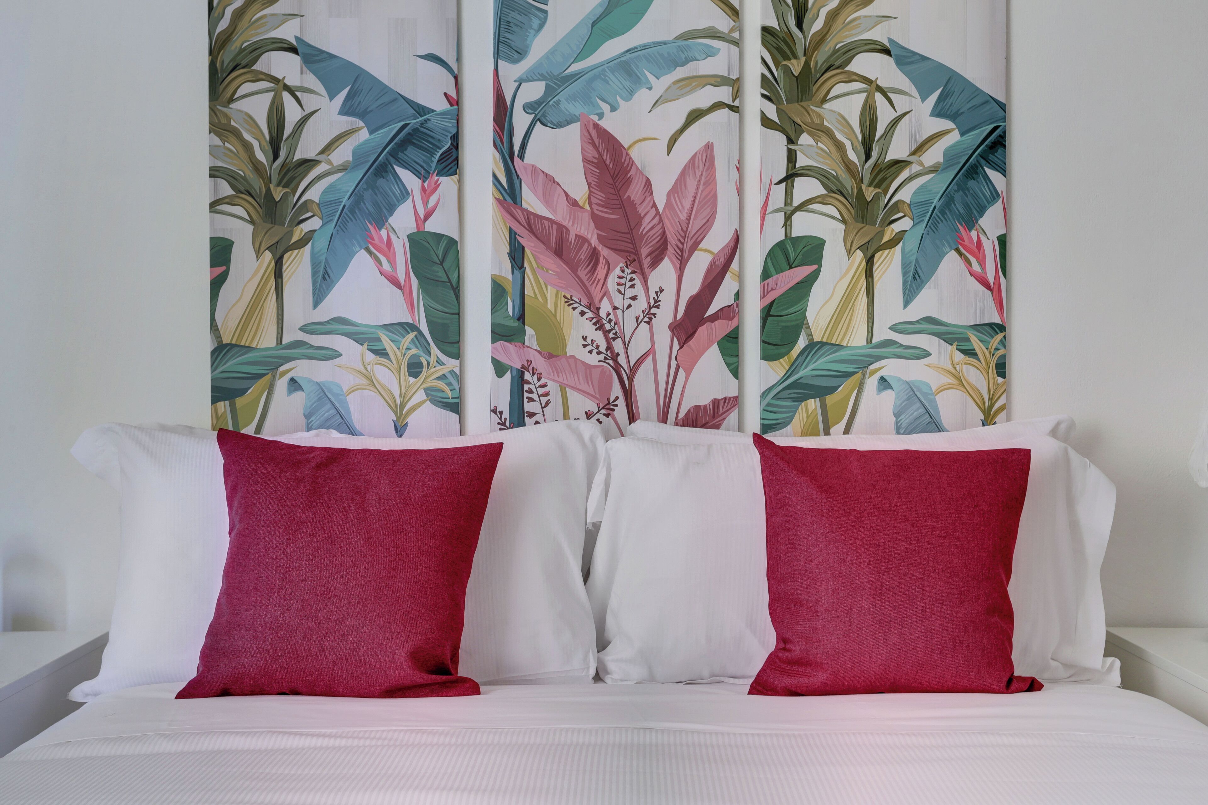 A bed with white bedding and two red pillows, set against a wall with a tropical leaf pattern.