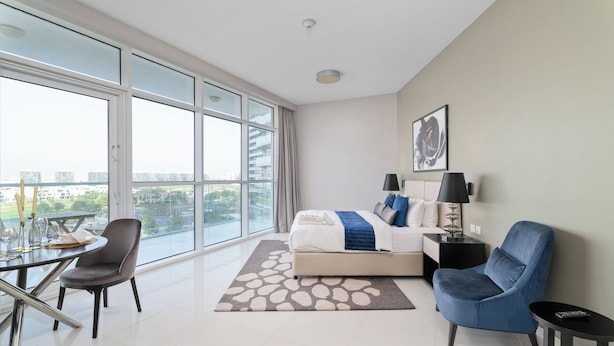 Een moderne hotelkamer met een ruim balkon, een bed met een blauw-witte dekbedovertrek, een grijze fauteuil, een kleine tafel met een glazen blad en een zwart nachtkastje met een lamp.