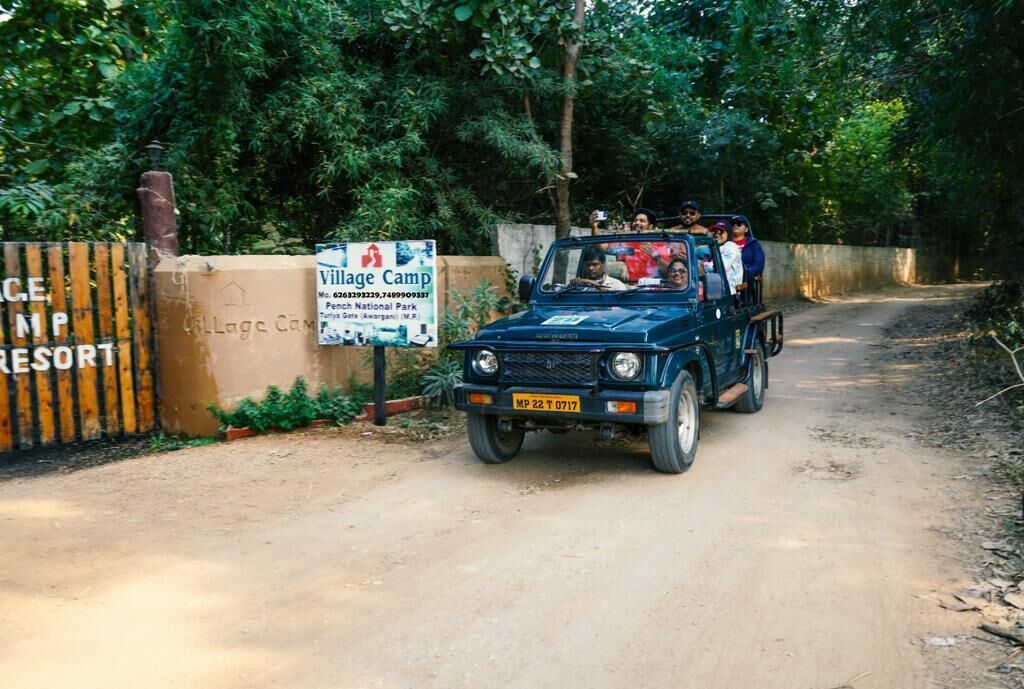 A blue vehicle with passengers on a dirt road, a sign for Village Camp, and a fence with partially visible text.
