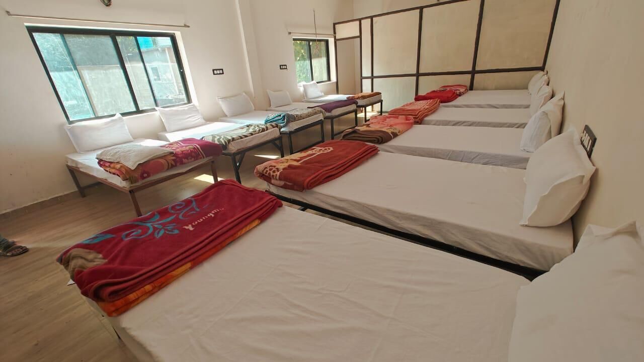 A dormitory room with four beds arranged in a row, a window providing natural light, and a small table between the beds.