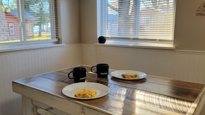 Habitación de esquina con dos ventanas, una mesa de madera con un plato de comida y una taza de café, y un altavoz en la pared.
