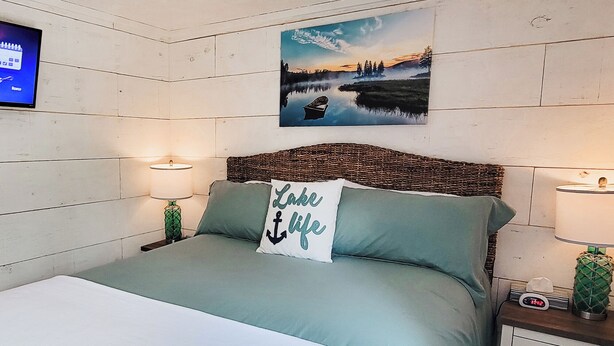 Un dormitorio con una cama, dos mesitas de noche, un televisor en la pared y un cuadro de un paisaje lacustre.