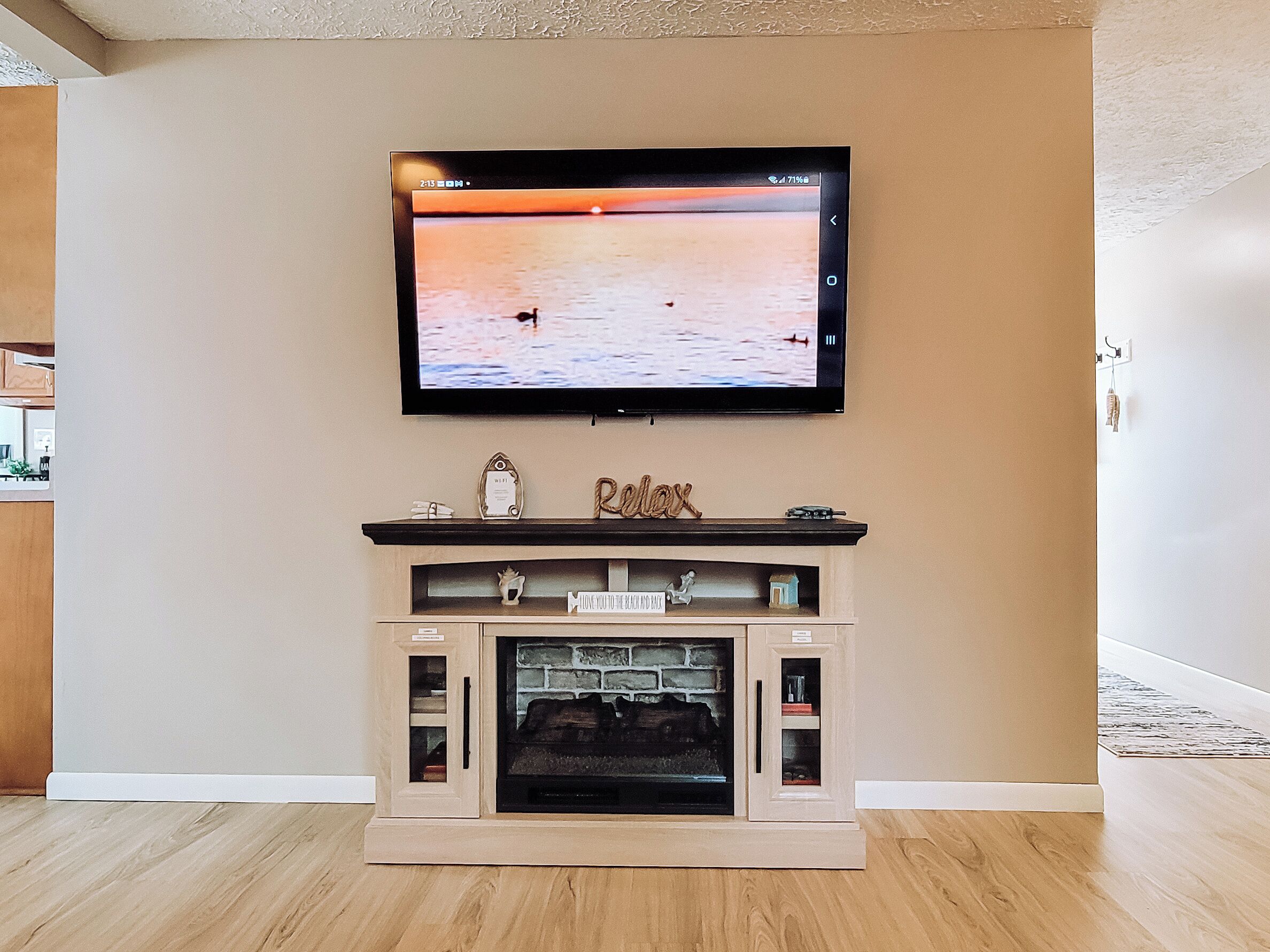 A living room with a flat-screen TV mounted above a fireplace, wooden flooring, and a beige wall.
