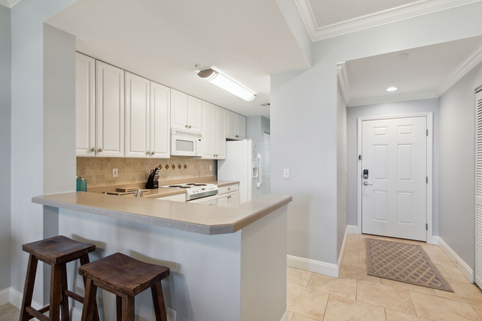 A kitchen with white cabinets, a countertop, and a microwave. There are two stools and a door leading to another room.