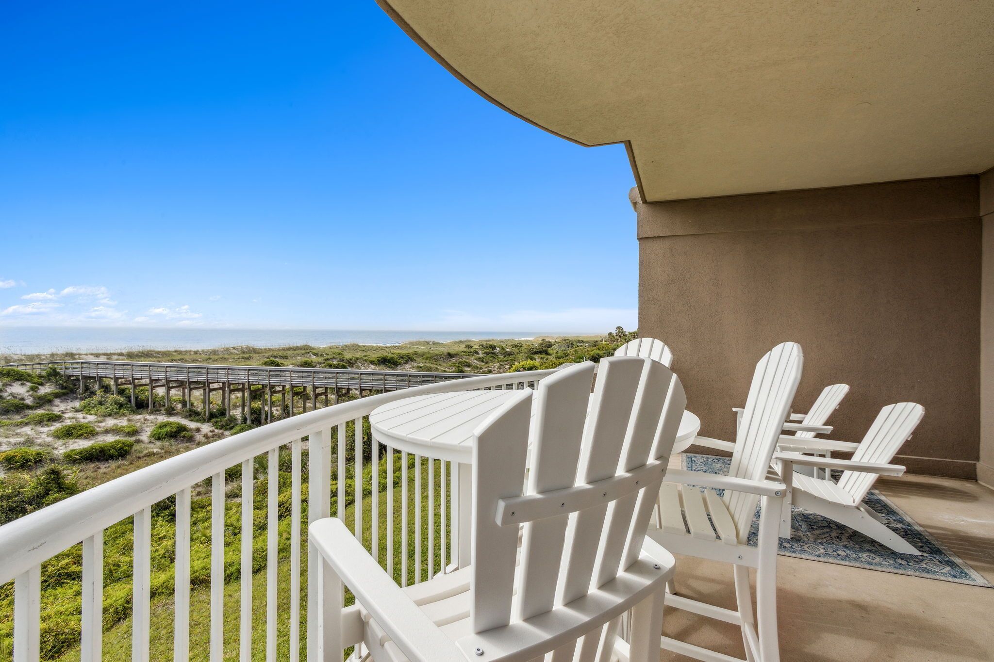 A balcony with white Adirondack chairs and a view of a beach and a bridge.