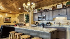 A kitchen with wooden cabinets, a central island with stools, a chandelier, and a dining area with a leather couch. - Mountain Views and City Lights- Convenient to Attractions-Hot Tub Game Tables (Pigeon Forge)