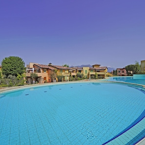 Uma grande piscina ao ar livre com deck ladrilhado, rodeada por edifícios com vários andares e varandas, e um céu azul claro.