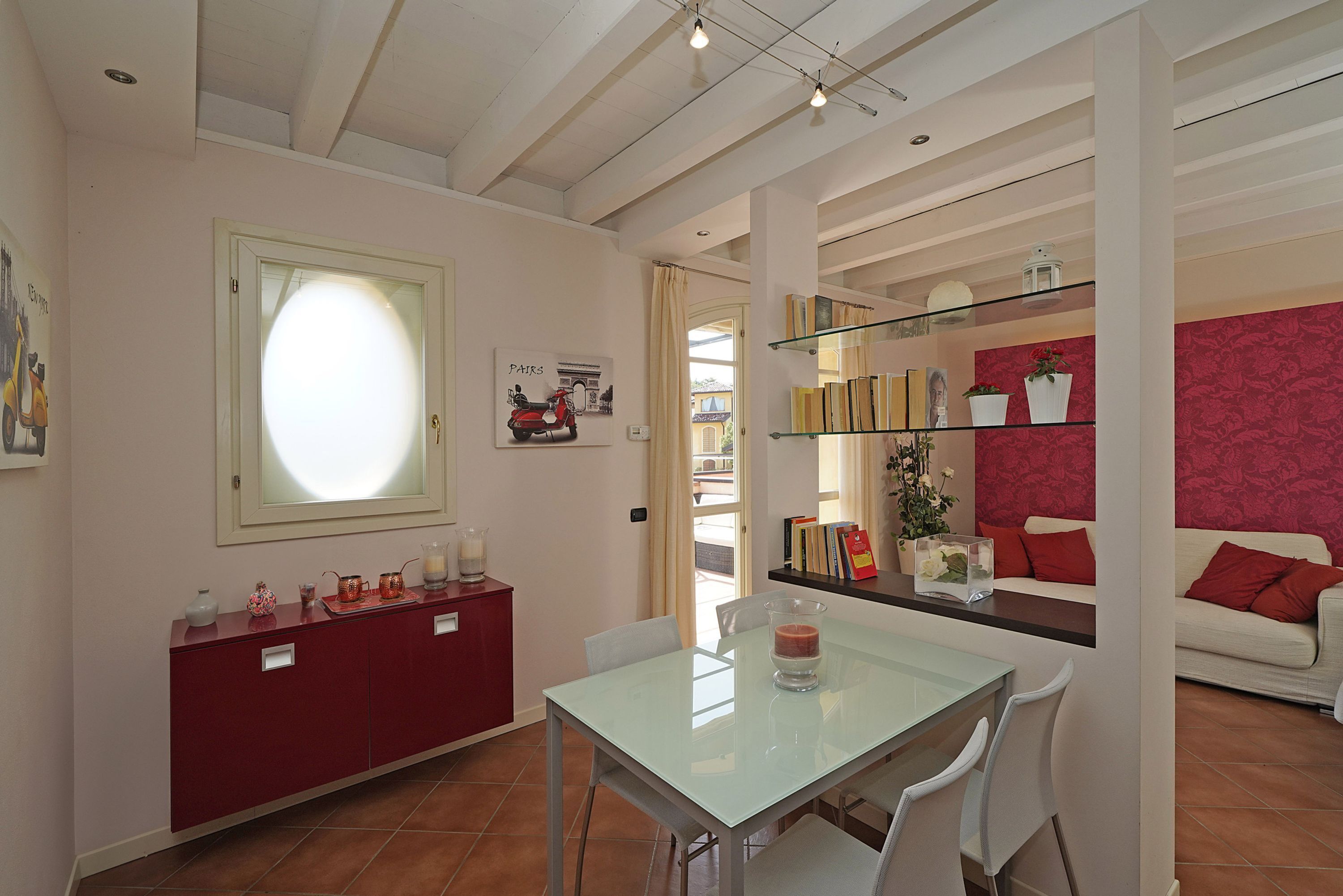 Un'area pranzo con un tavolo in vetro e sedie, un armadietto rosso con oggetti decorativi, una libreria con libri e un vaso, e una parete con motivo rosso.