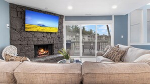 A living room with a stone fireplace, a large flat-screen TV, and a view of a residential area through large windows. - Our Cove Retreat - Perfect for Wave-Free Swimming, Tanning & Prime Location Fun! (Lake Ozark)