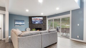 A living room with a sectional sofa, a stone fireplace, a flat-screen TV, and a sliding glass door leading to an outdoor area. - Our Cove Retreat - Perfect for Wave-Free Swimming, Tanning & Prime Location Fun! (Lake Ozark)