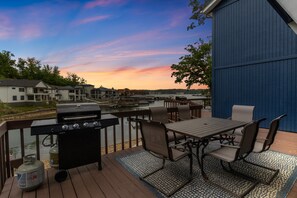A deck with a grill, outdoor table, chairs, and a view of a body of water and buildings. - Our Cove Retreat - Perfect for Wave-Free Swimming, Tanning & Prime Location Fun! (Lake Ozark)