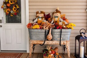 A rustic outdoor setting with a metal table holding autumn decorations, two scarecrow figures, and a welcome mat. - Peaceful Country Stay Covered Porch, Fire-pit and Wildlife (Mendota)