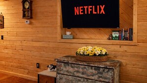 Ein gemütliches Zimmer mit einer Holzwand, einem Fernseher, der “NETFLIX” zeigt, einer Uhr, einem kleinen Regal mit Büchern und einem Teddybären sowie einem Blumenkorb auf einem Schminktisch.
