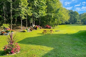 A grassy area with a picnic table, surrounded by trees and colorful flower beds. - Peaceful Country Stay Covered Porch, Fire-pit and Wildlife (Mendota)