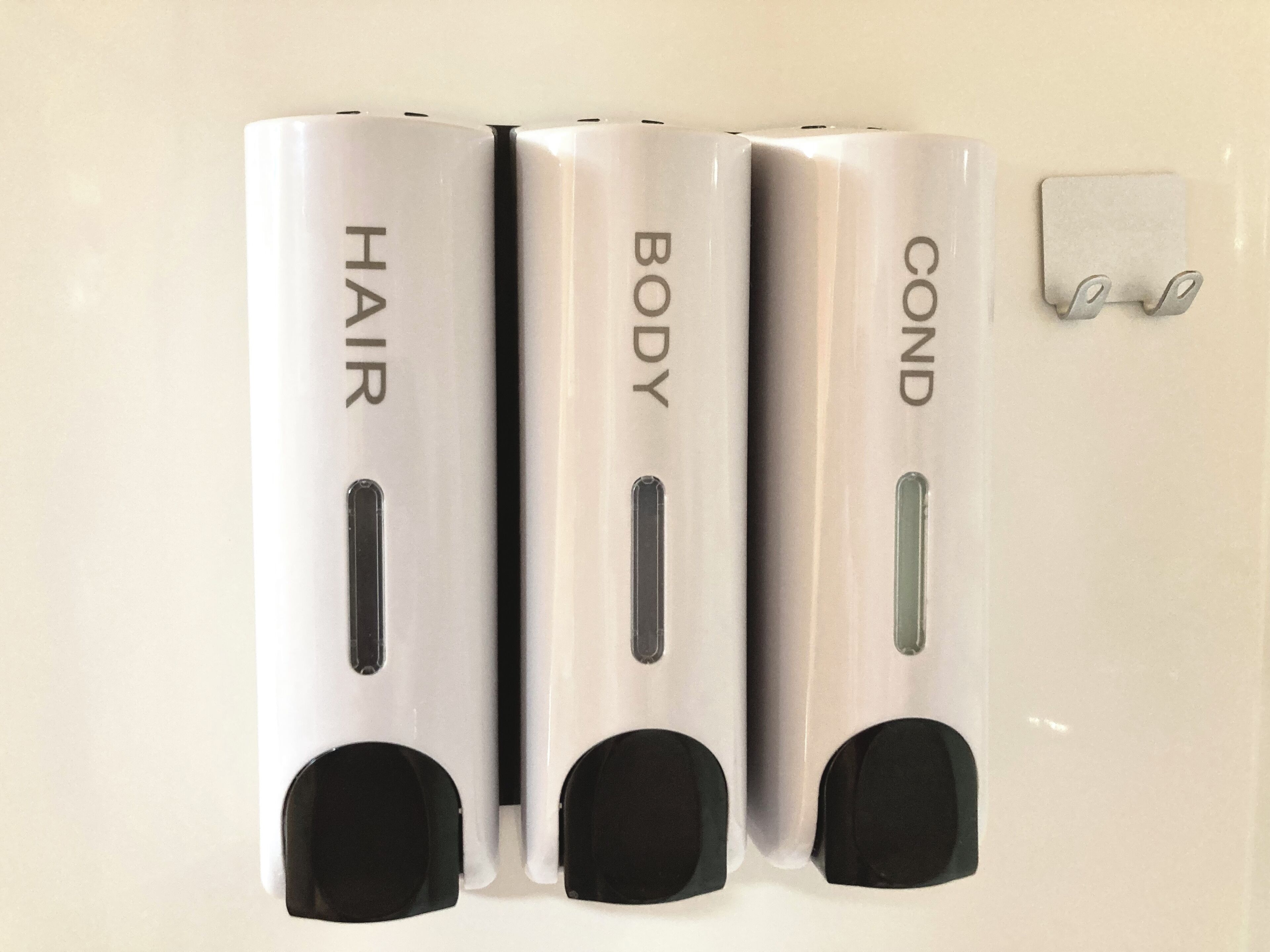Drei wandmontierte Spender mit den Beschriftungen 'HAIR', 'BODY' und 'COND' sowie ein kleiner Haken an der Wand.