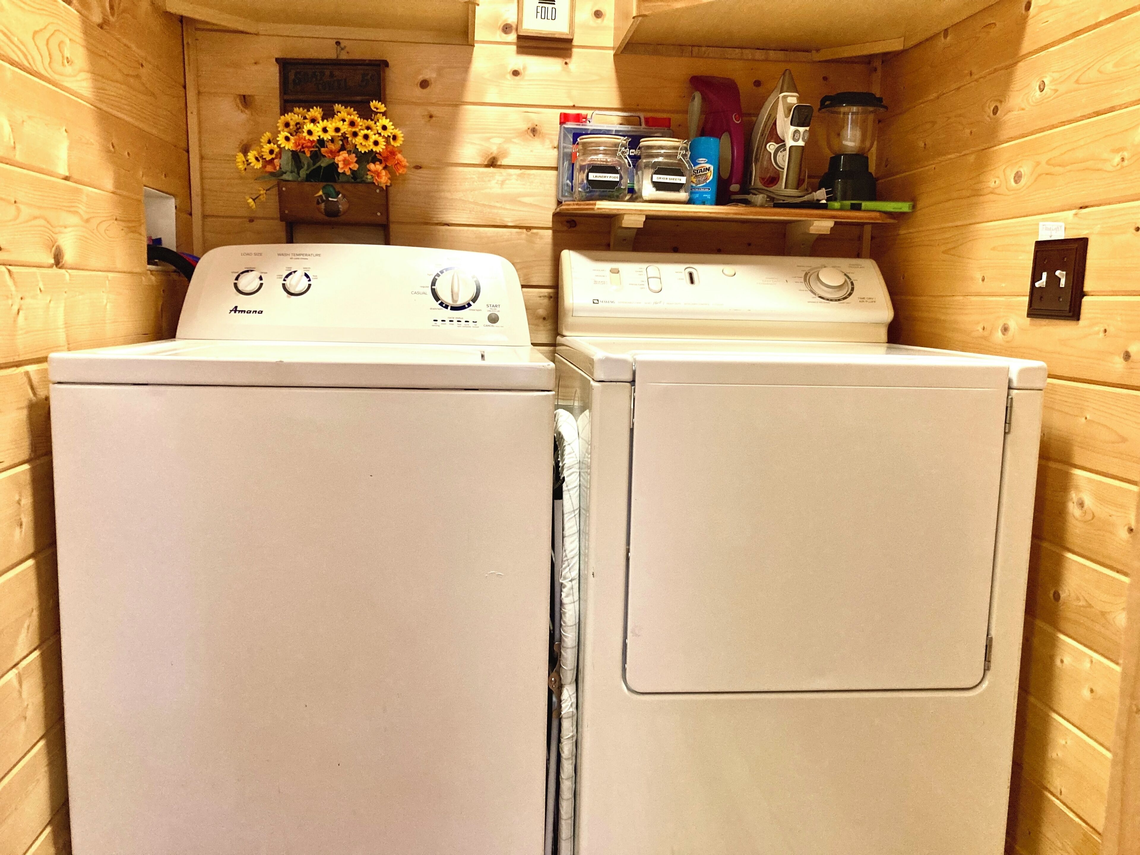 A laundry room with two white front-loading washers and dryers, wooden walls, a shelf with cleaning supplies, and a vase of sunflowers.