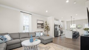 Ein modernes Wohnzimmer mit einem grauen Sofa, einem runden Couchtisch und einer Küchenzeile mit weißen Schränken und einem schwarzen Kühlschrank.