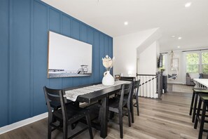 A dining area with a dark wooden table, chairs, and a white vase with dried flowers. A large framed picture of a beach scene is on the wall.
