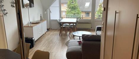 Et moderne køkken med hvide skabe, et spiseområde med et rundt bord og stole, og en opholdsstue med en sofa og et fjernsyn.