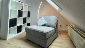 A loft bedroom with a sloped ceiling, a built-in white shelving unit, a grey sofa with a pillow, and a radiator on the floor. - 1 bedroom apartment in grã¶mitz (Grömitz)