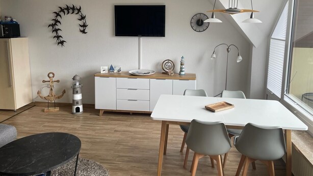 A modern living room with a white dining table, grey chairs, a black sofa, a TV mounted on the wall, and a wooden cabinet. - 1 bedroom apartment in grã¶mitz (Grömitz)