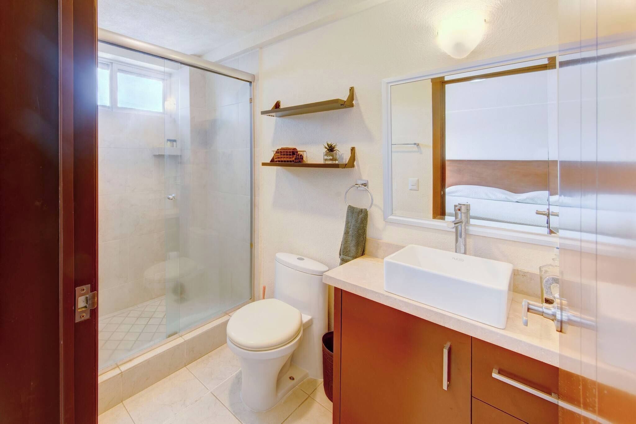Een badkamer met een glazen douchecabine, toilet, wastafel met spiegel en houten planken boven het toilet.