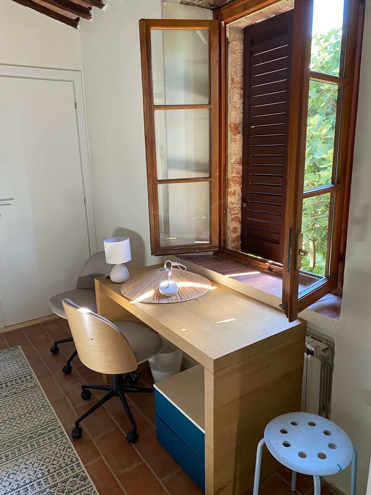 Un petit espace de travail comprenant un bureau en bois, une chaise, un tabouret et une fenêtre avec des volets en bois.