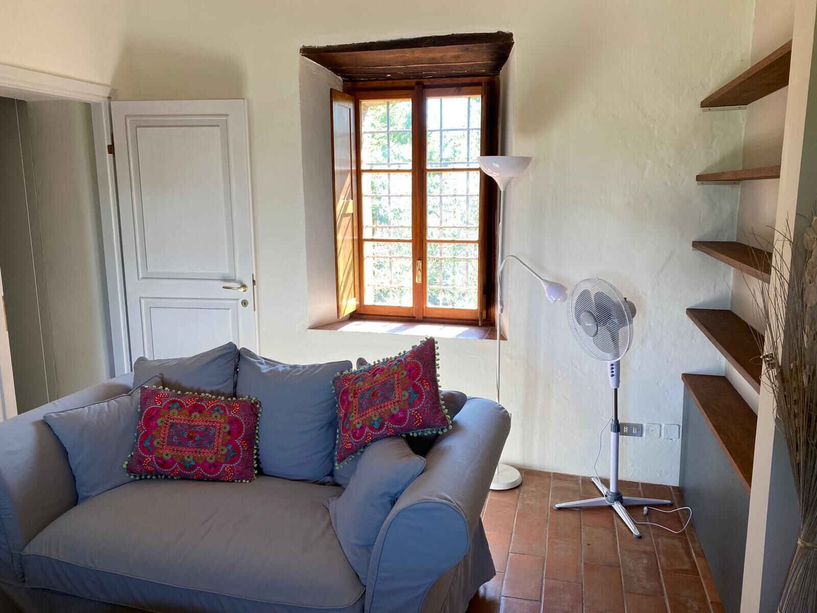A room with a light blue sofa, two colorful pillows, a white door, a window with a wooden frame, a standing fan, and a set of wooden shelves.