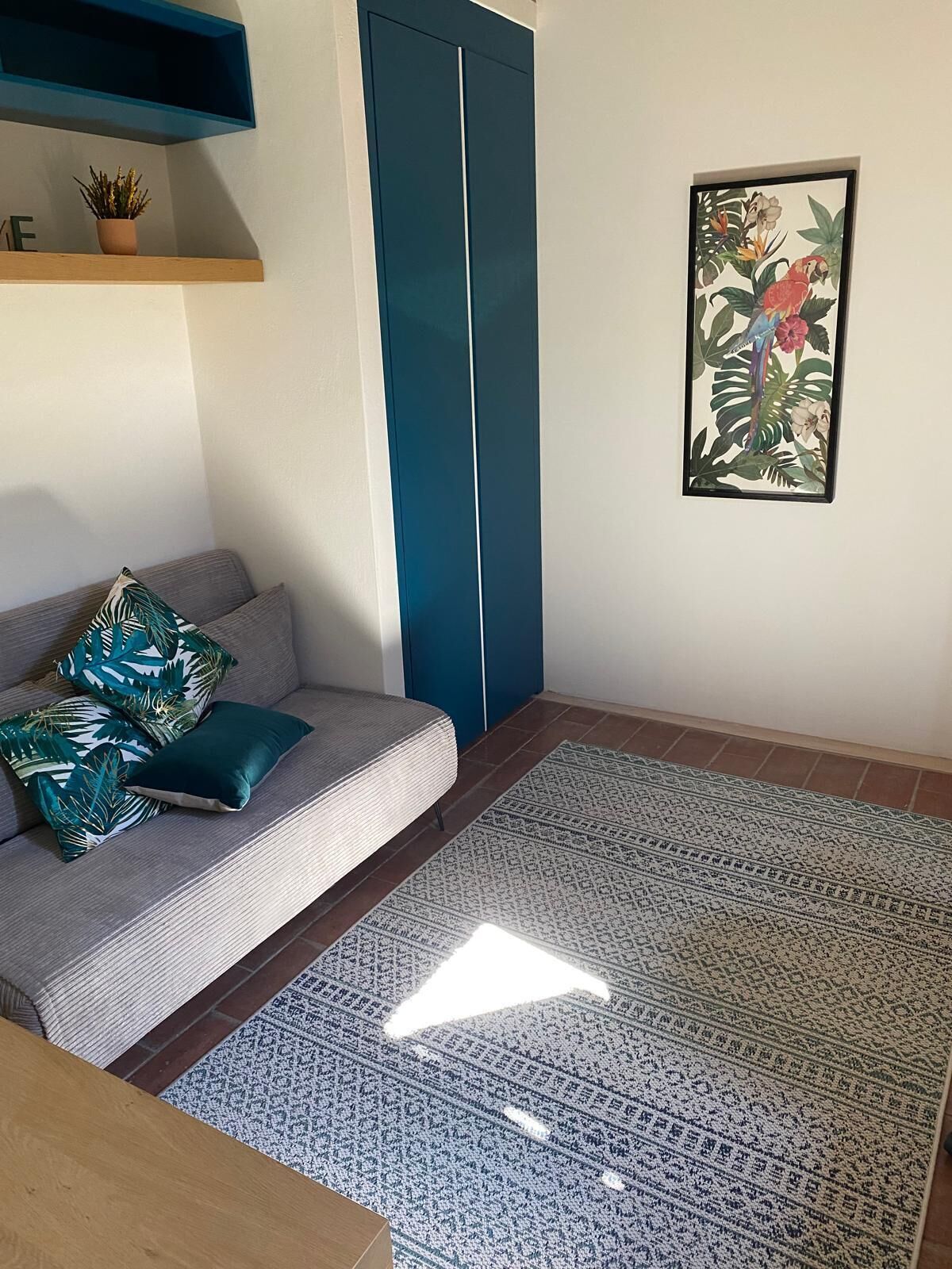 A living room with a grey sofa, patterned cushions, a blue accent wall, a framed tropical artwork, and a patterned rug.