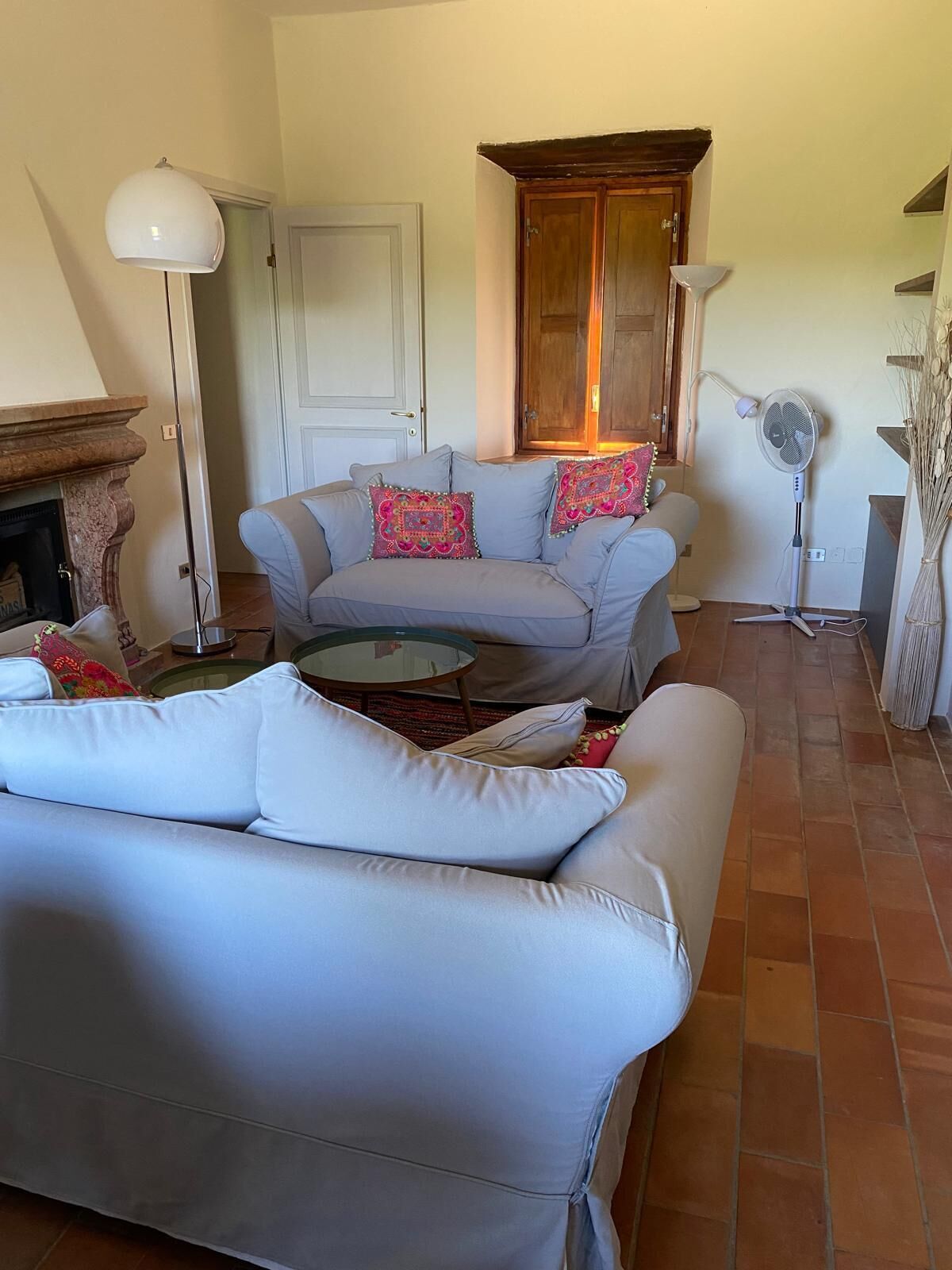 Un salon avec un canapé, un fauteuil et une petite table. On y trouve une cheminée, un ventilateur sur pied et une porte au fond.