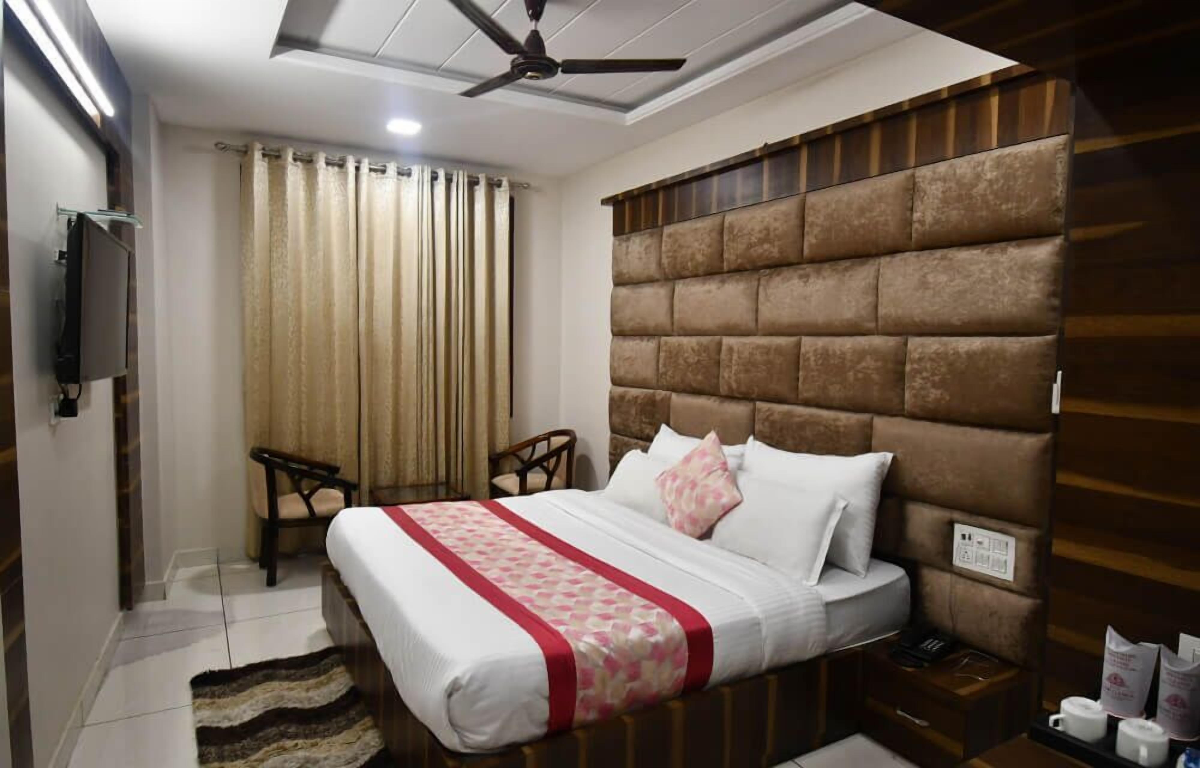 A hotel room with a bed, a ceiling fan, a TV, a window with curtains, and a wooden headboard.