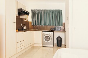 Compact kitchen with washing machine, stainless steel kettle, and trash bin.