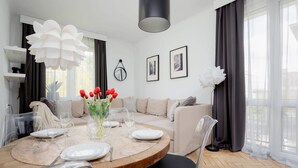 A modern living room with a round dining table set for four, a beige sofa, and a large white flower chandelier. - Stylish Apartment with Balcony | Close to Zoo and Workspace (Warszawa)