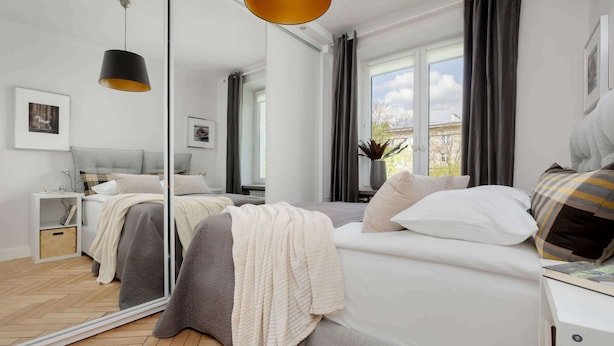A modern bedroom with a large mirror, a bed with white and gray bedding, a bedside table, and a window with curtains. - Stylish Apartment with Balcony | Close to Zoo and Workspace (Warszawa)