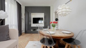 A modern living room with a round wooden dining table, clear plastic chairs, a sofa, and a TV mounted on the wall. - Stylish Apartment with Balcony | Close to Zoo and Workspace (Warszawa)